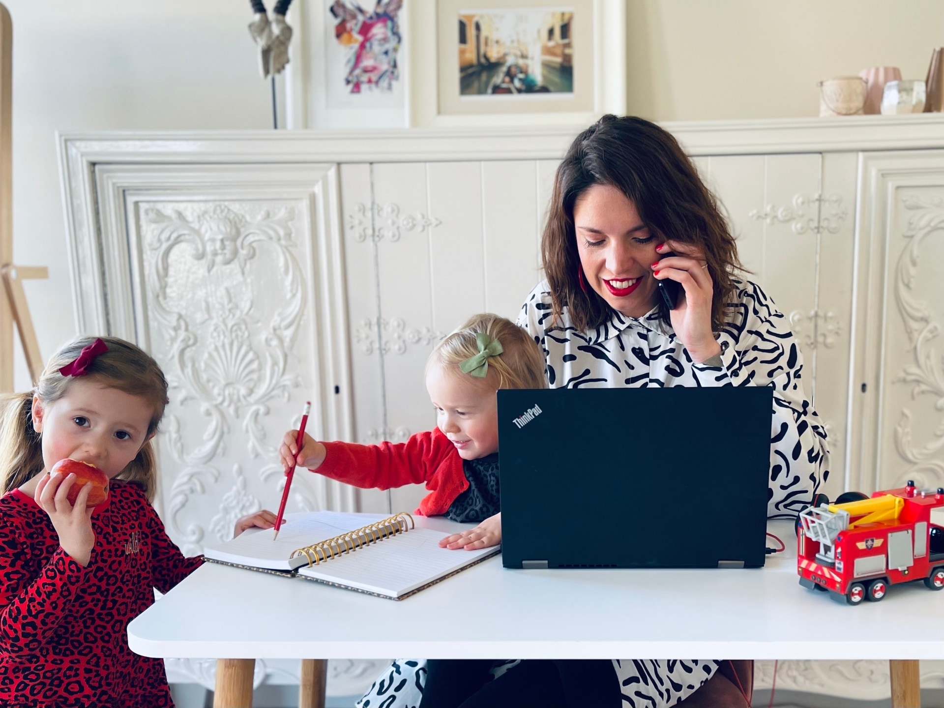 A woman on the phone whilst sat at her desk on her laptop with two children in the picture