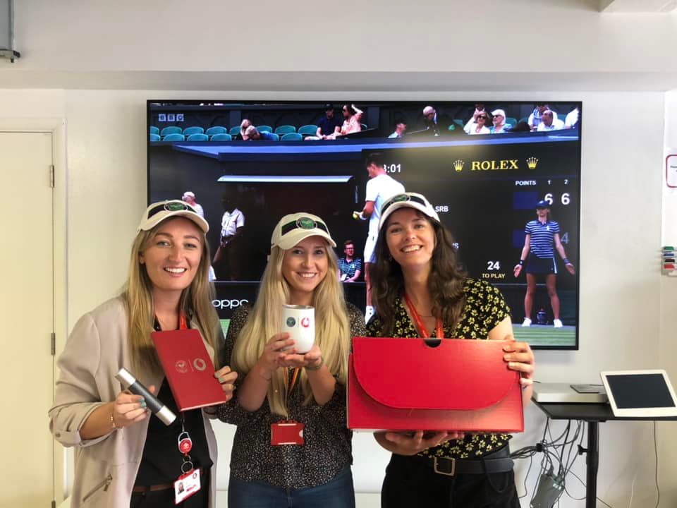 Three women standing in front of a screen showing tennis with Wimbledon hats on
