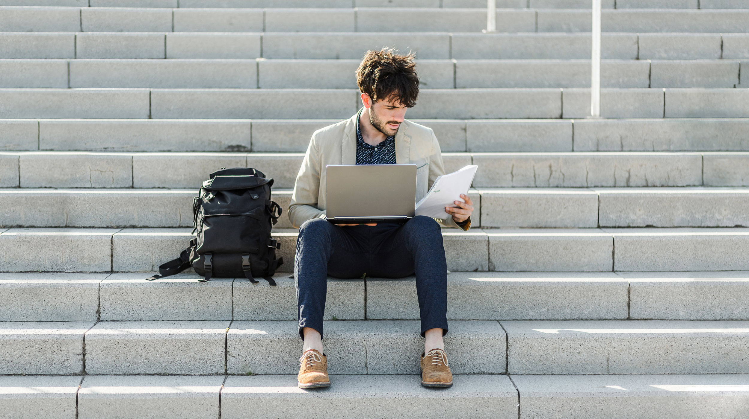 A man sat on steps outside holding a laptop and a piece of paper