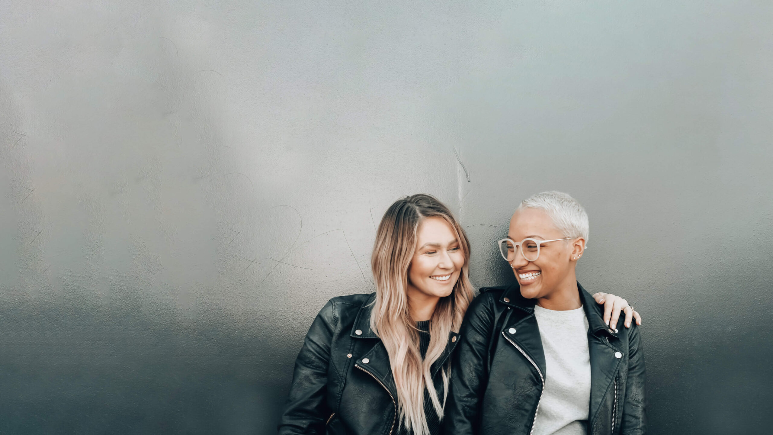 Two people smiling at each other with one having their arm around the other against a plain grey background