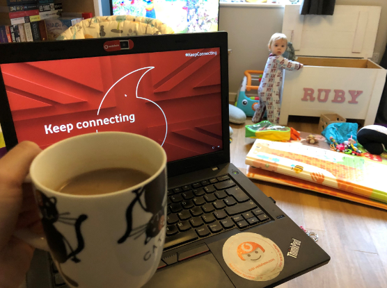 A image of a person holding a cup of tea in front of a laptop with their child in the background playing
