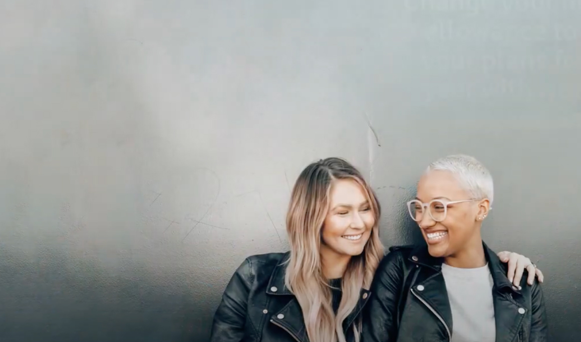 Two people smiling at each other with one having their arm around the other against a plain grey background