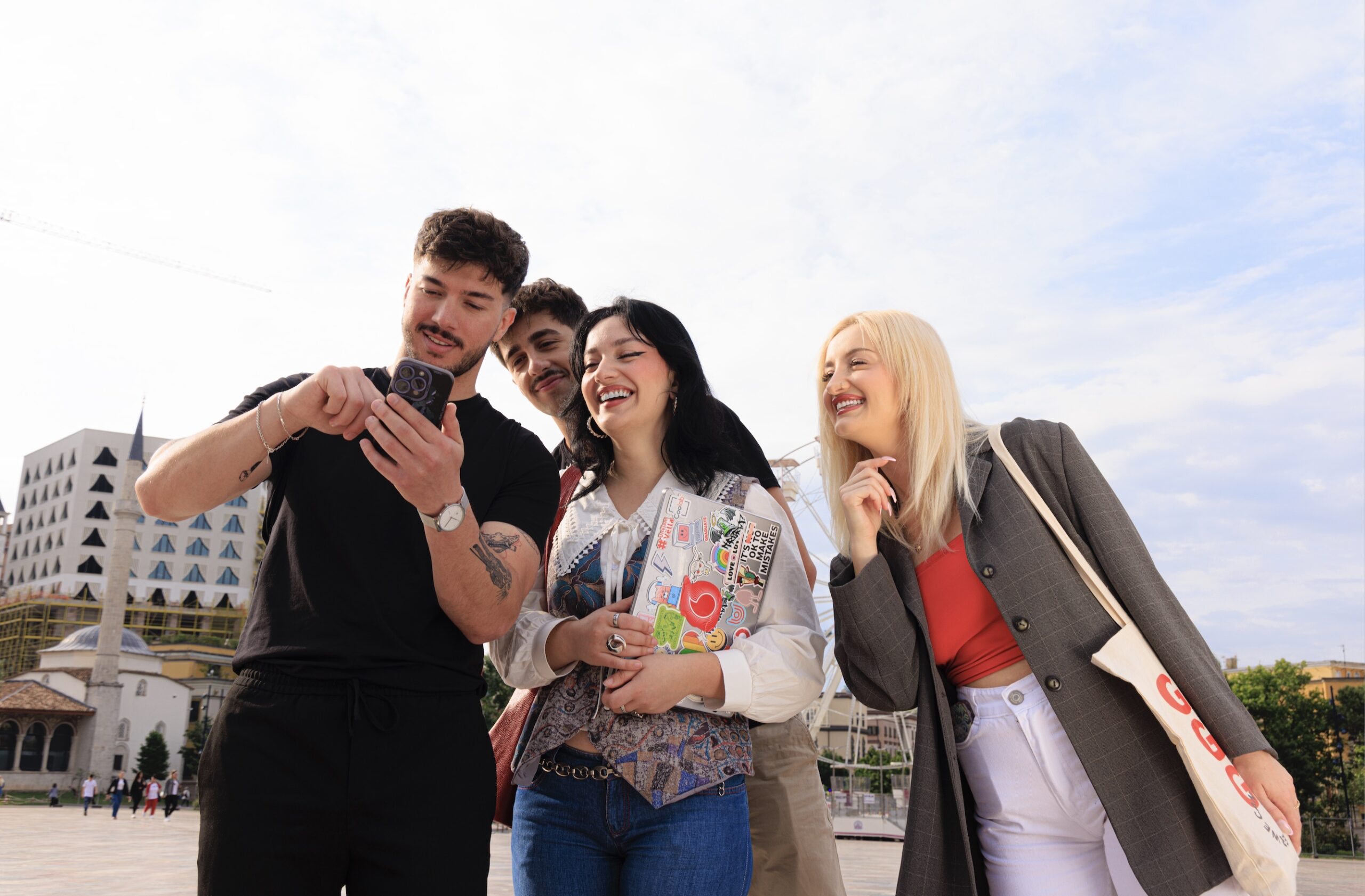 Group of two males and two females are outside gathering around a phone to see the screen