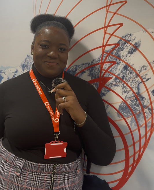 woman stood Infront of a Vodafone sign holding a small mic in black long Sleeve top