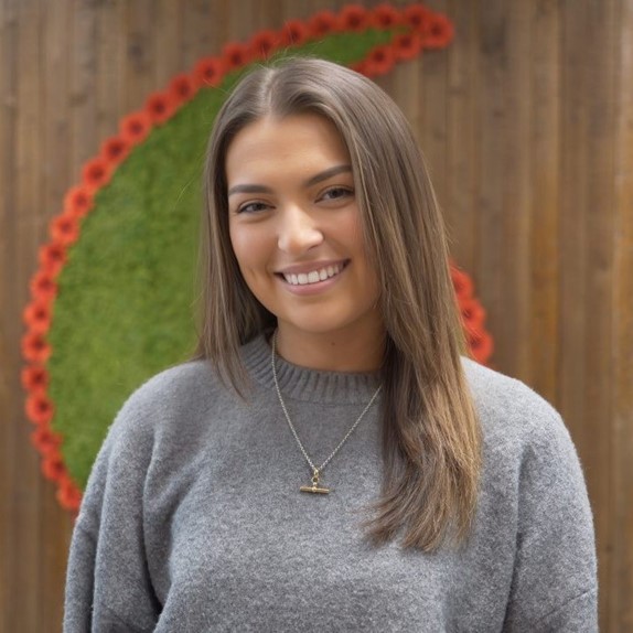 Brunette woman stood Infront of a Vodafone logo in a grey jumper.