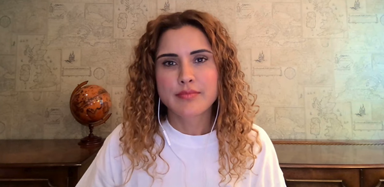 Woman speaking to camera during a video recording, wearing a white top and earphones, seated indoors with a globe visible in the background.